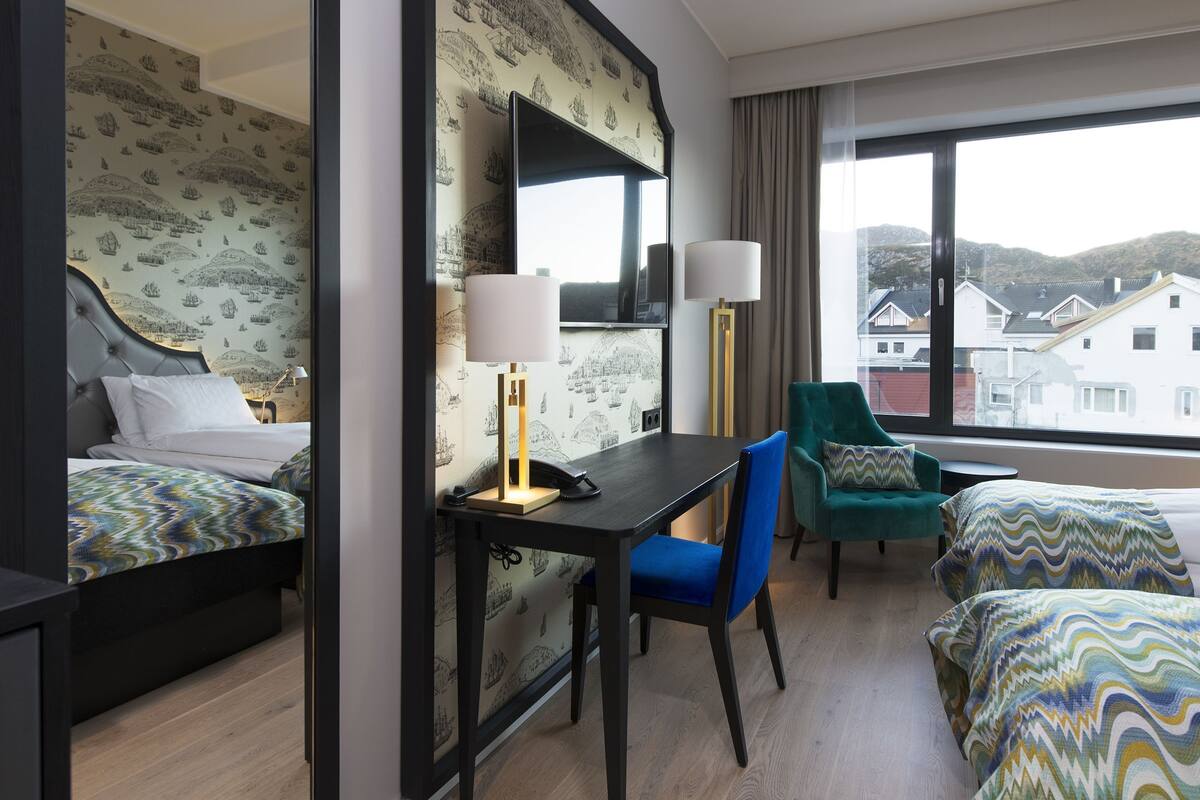 Quarto de hotel com duas camas, uma escrivaninha com abajur, uma cadeira e uma janela grande com vista para edifícios.