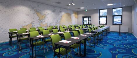 Een vergaderzaal met groene stoelen, een grote kaart aan de muur en een projectiescherm.