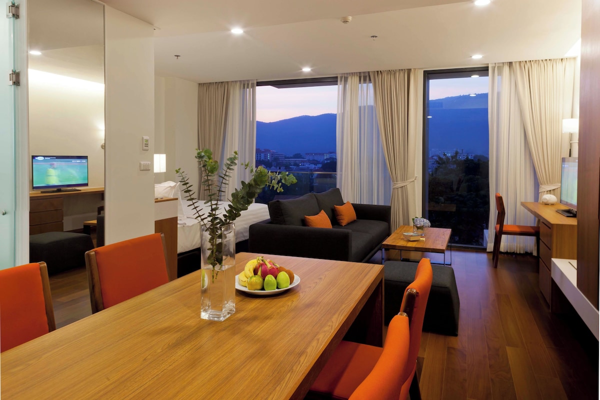 A modern hotel room with a dining area, a sofa, a TV, and a view of the mountains.