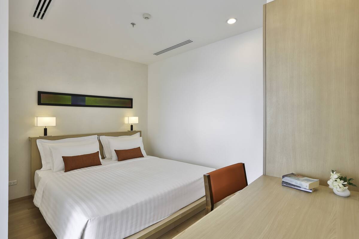 A hotel room with a bed, bedside lamps, a small table with books and a vase of flowers, and a wooden headboard.