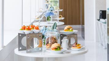 Arredamento moderno con un tavolo bianco su cui sono esposti cibi e bevande, un supporto metallico con fiori e una parete rivestita in legno sullo sfondo.