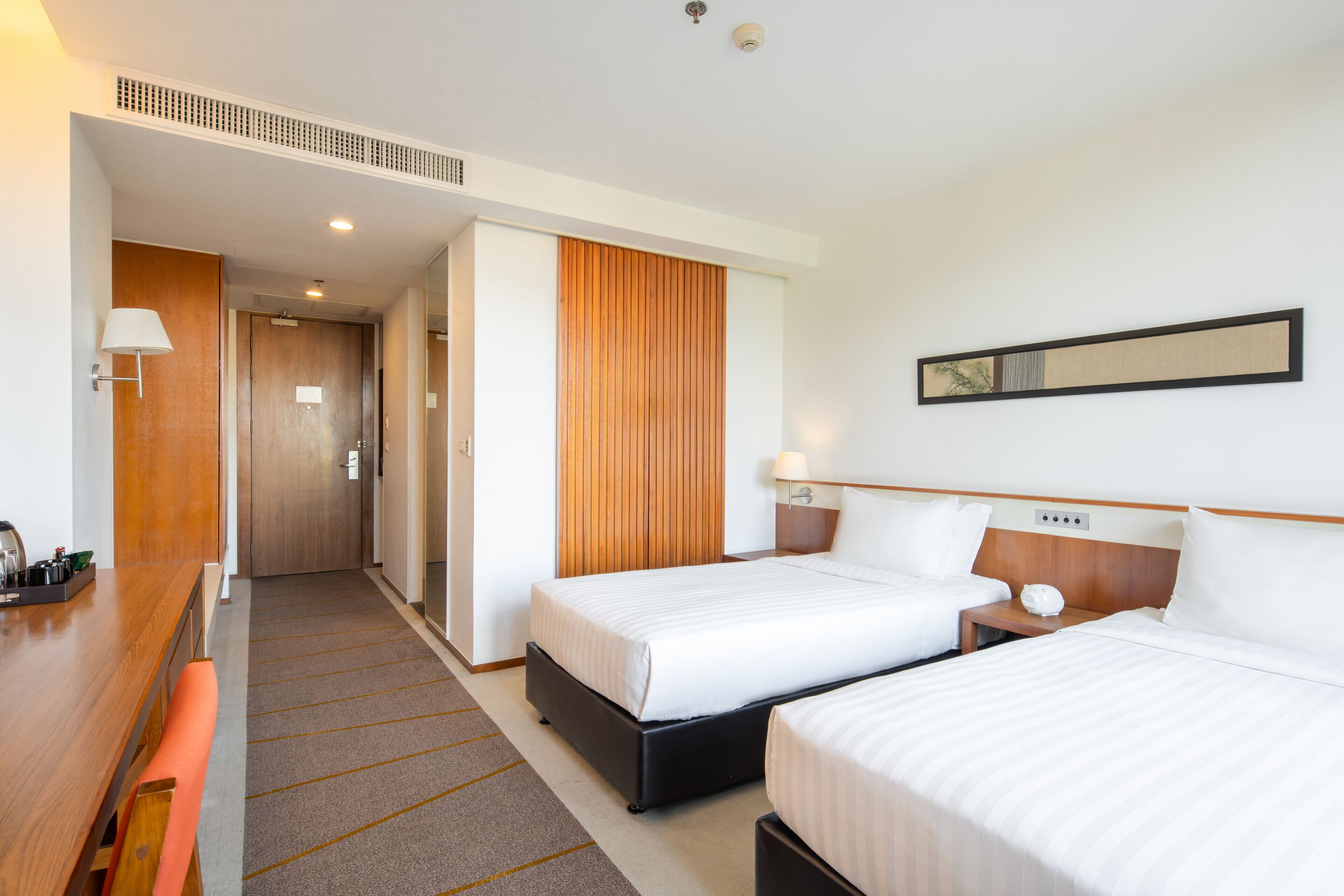 A hotel room with two beds, a wooden desk, a bench, a wall-mounted lamp, and a door leading to a hallway.