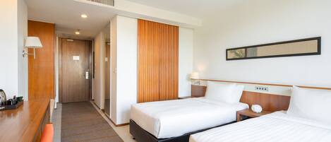 A hotel room with two beds, a wooden desk, a bench, a wall-mounted lamp, and a door leading to a hallway.