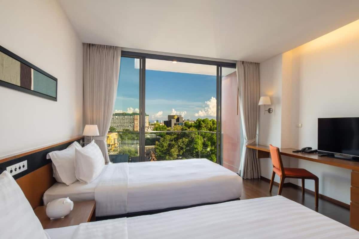 A hotel room with two beds, a desk, a chair, a TV, and large windows with a city view.
