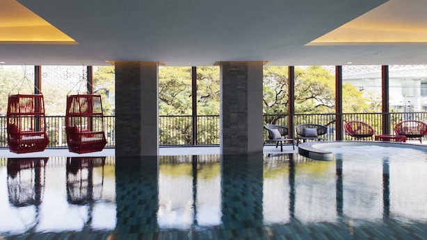 Una piscina interior amplia con paredes de vidrio transparente, sillas de mimbre rojas y vistas a árboles.