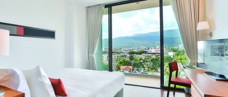 A hotel room with a large bed, a desk with a computer, and a view of a cityscape through a large window.