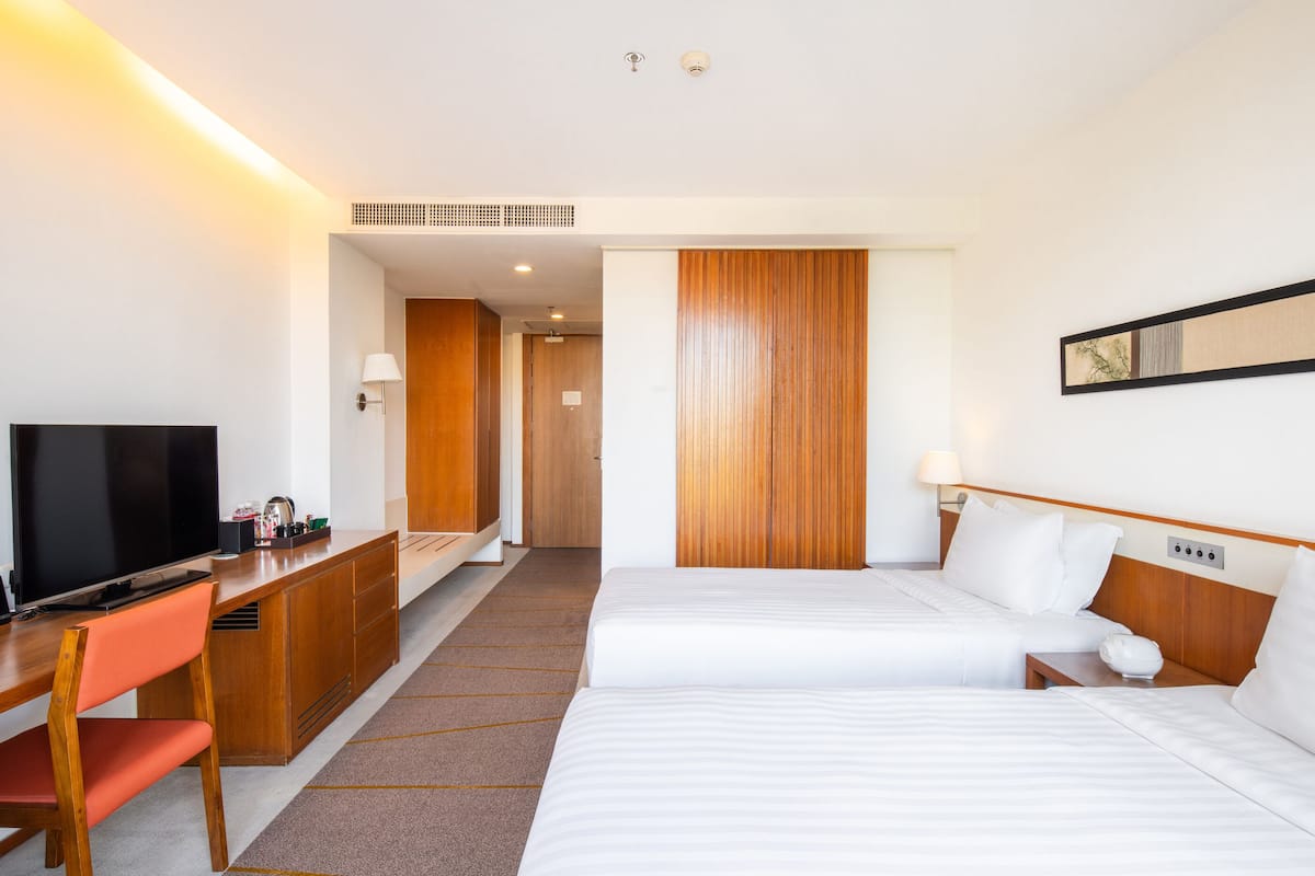 A hotel room with two beds, a desk with a TV, a chair, a wooden panel wall, and a framed picture on the wall.