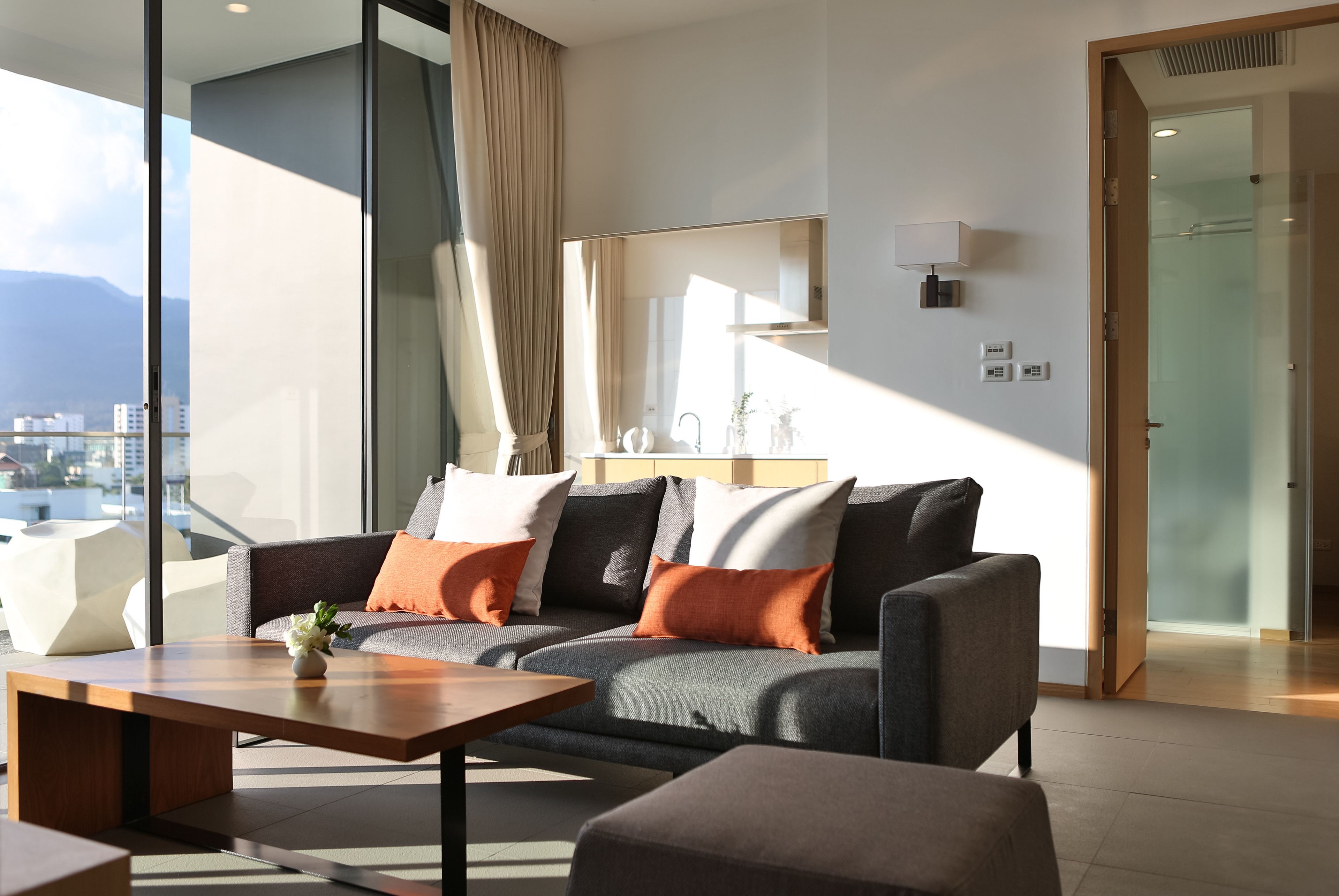 A modern living room with a grey sofa, orange cushions, a wooden coffee table, and a large window offering a city view.