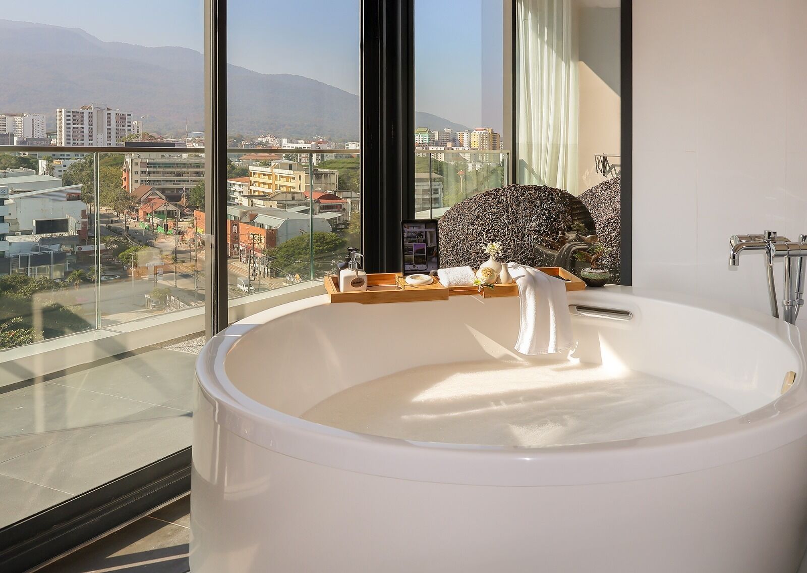 A modern bathroom with a large bathtub, a wooden tray with toiletries, and a city view through large windows.