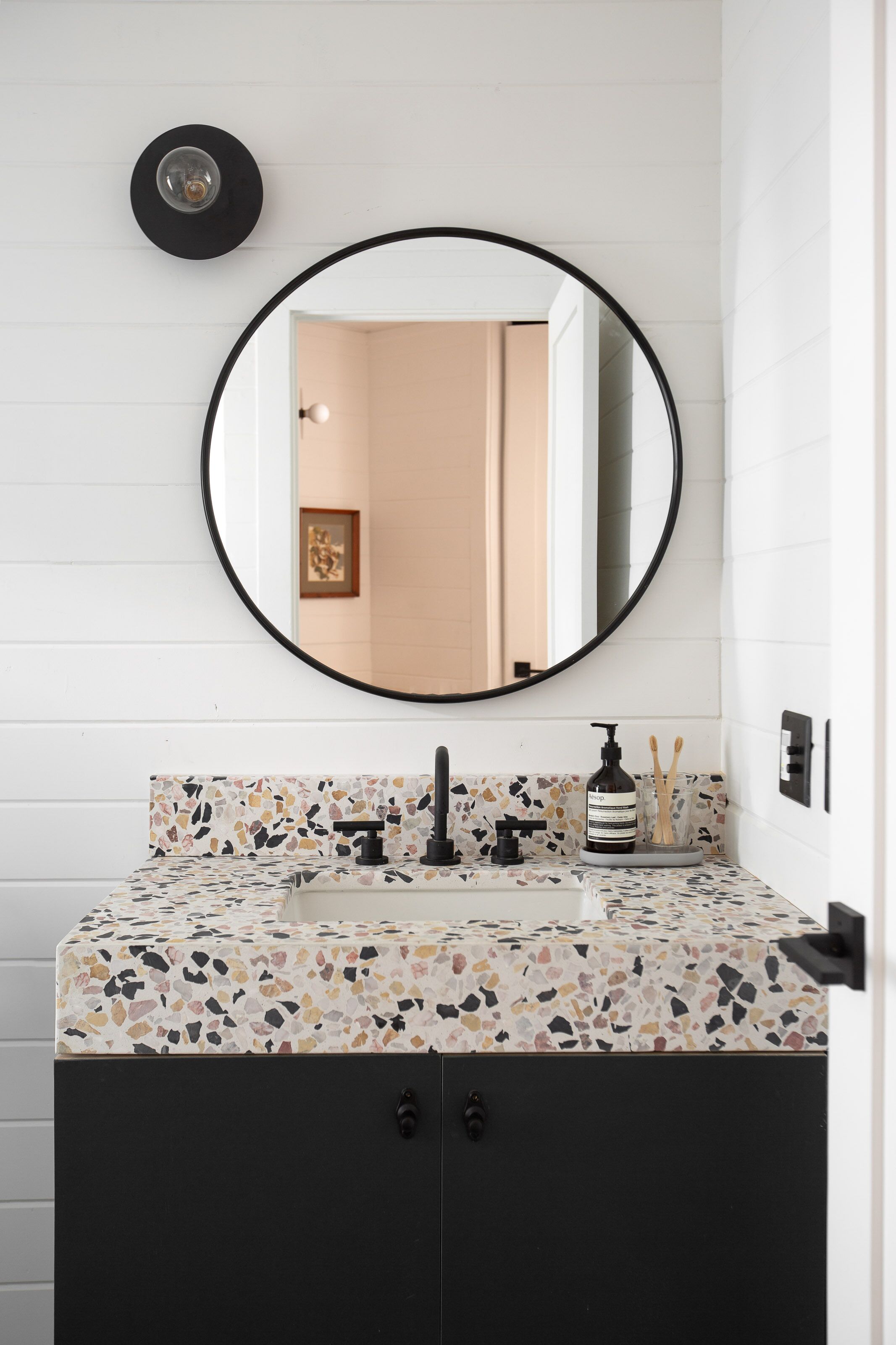 Een badkamer met een ronde spiegel, een zwarte kraan, een zeepdispenser en een tandenborstelhouders.