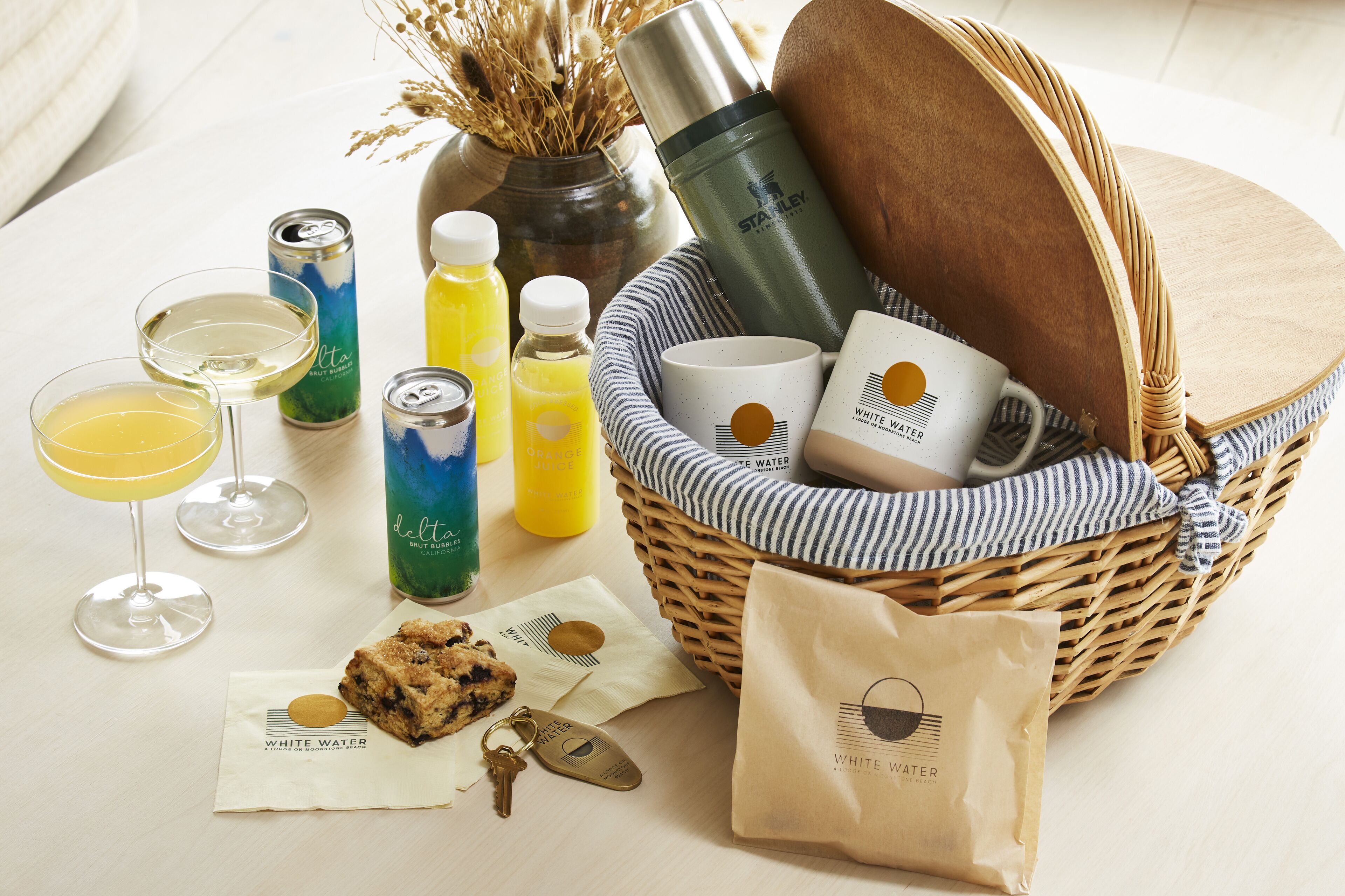 Ein Korbwicker-Korb mit einer Edelstahl-Wasserflasche, einer Tasse, einer Dose, einer Saftflasche, einem Glas Wein, einem Keks, einem Servietten und einem Schlüsselanhänger, sowie einem Papierbeutel mit dem White Water Logo.