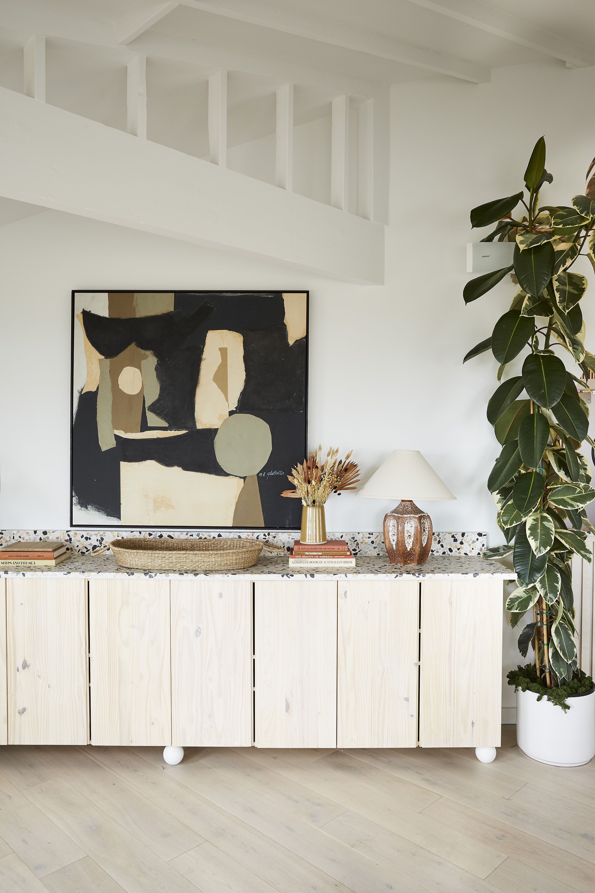 A modern living room with a wooden cabinet, a large abstract painting, a potted plant, and a lamp.