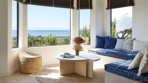A room with a curved white table, a woven chair, a blue sofa with cushions, and large windows overlooking the ocean. - White Water (Cambria)