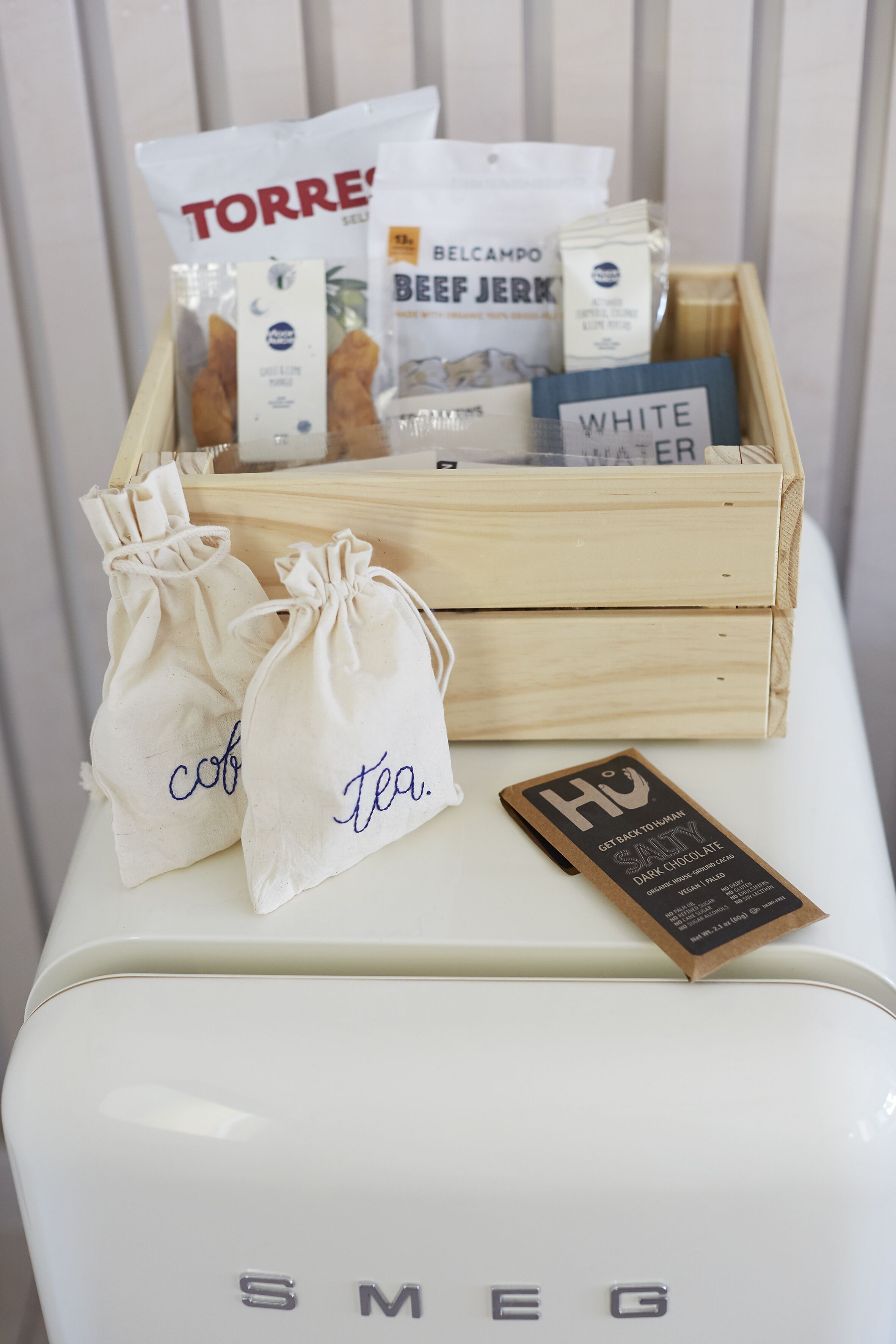A white Smeg refrigerator with a wooden crate containing various food items and two drawstring bags labeled "cob" and "tea."