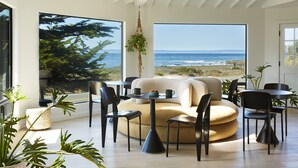 A modern living room with a large window overlooking the ocean, a round sofa, and a dining area with chairs and tables. - White Water (Cambria)