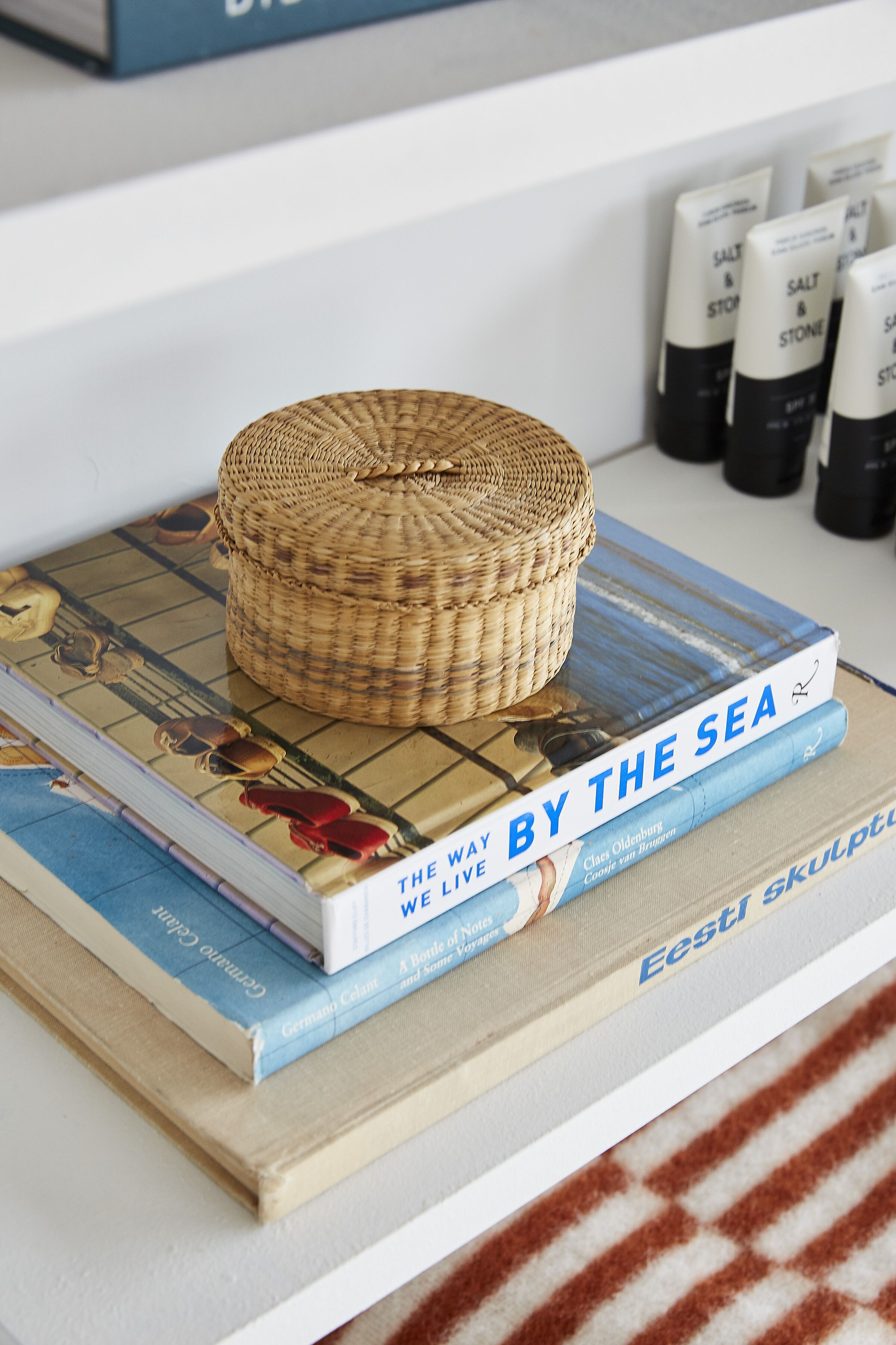 Una pila de libros con una cesta tejida encima, colocada sobre una estantería y una toalla a rayas debajo.