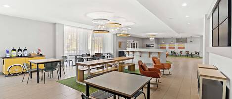 A modern dining area with multiple tables and chairs, a yellow counter with beverages, and a kitchen in the background.