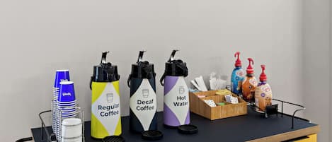 Estación de café con dispensadores etiquetados para café normal, café descafeinado y agua caliente, además de condimentos y servilletas.