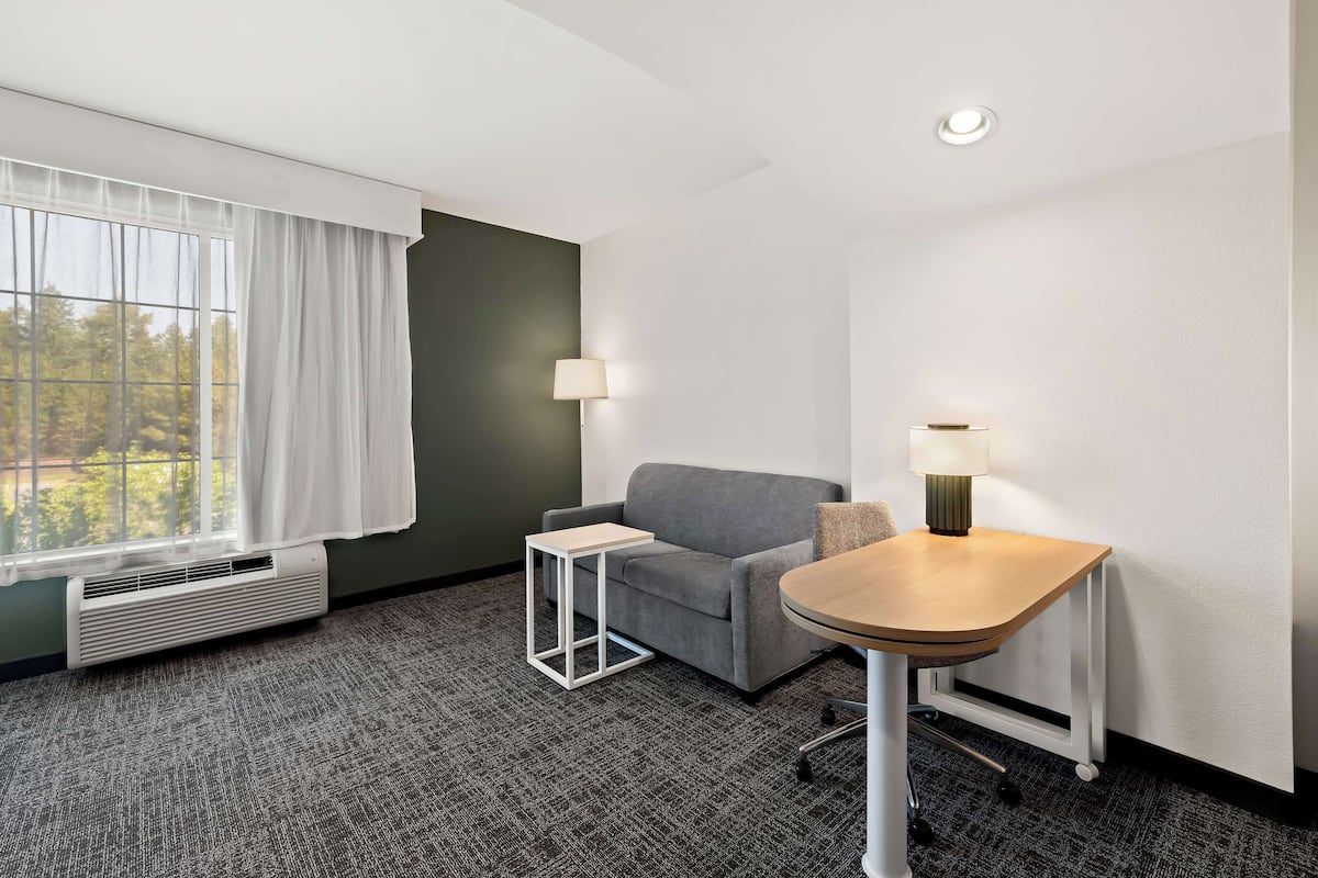 A hotel room with a gray sofa, a small white table, a lamp, and a window with white curtains.
