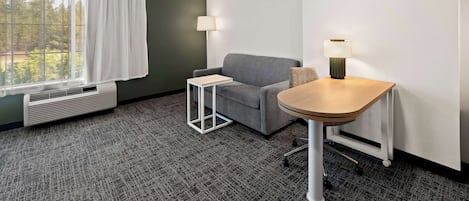 A hotel room with a gray sofa, a small white table, a lamp, and a window with white curtains.
