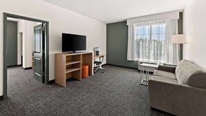 A hotel room with a grey carpet, a grey sofa, a TV on a wooden stand, a desk with a chair, and a window with sheer curtains.