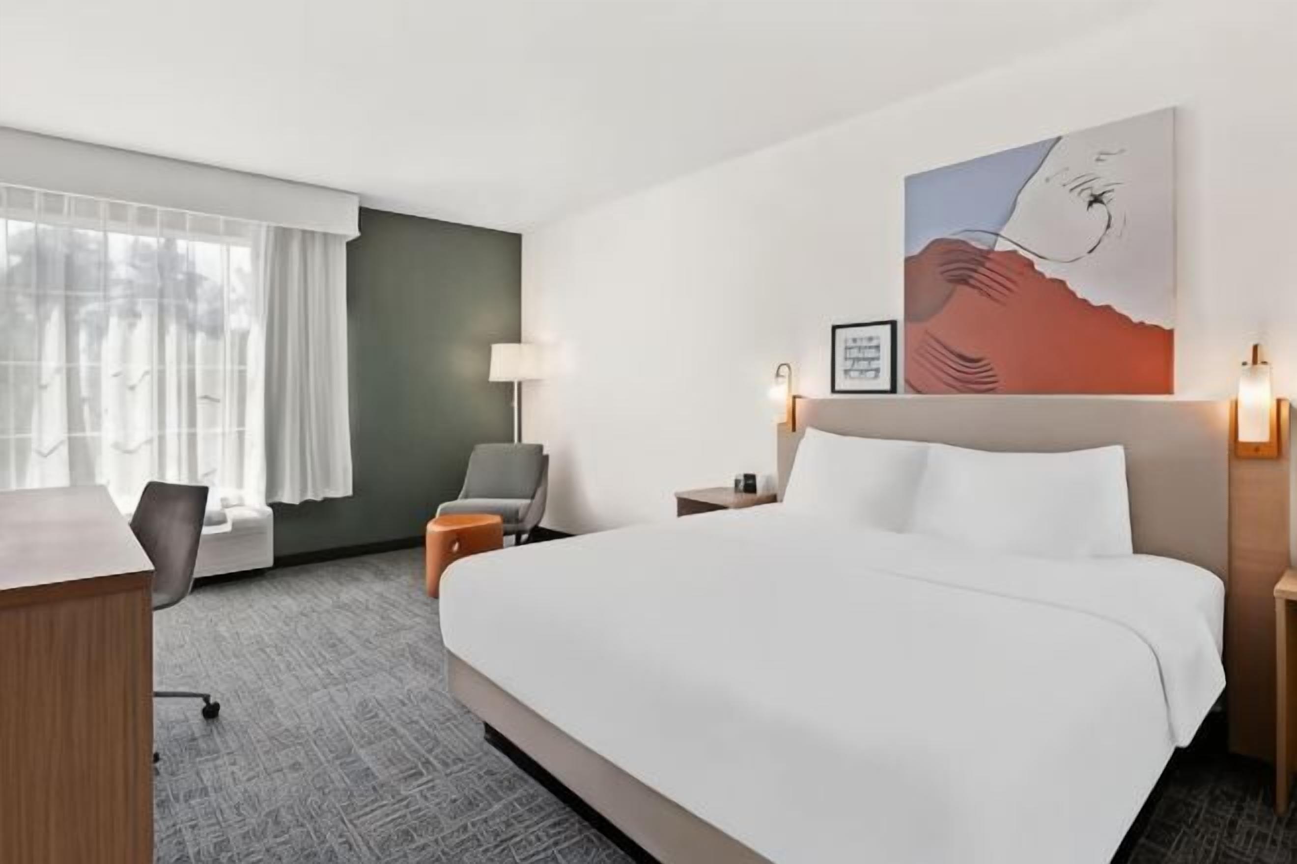 A hotel room with a large bed, a desk, a chair, a small table, and a window with curtains.