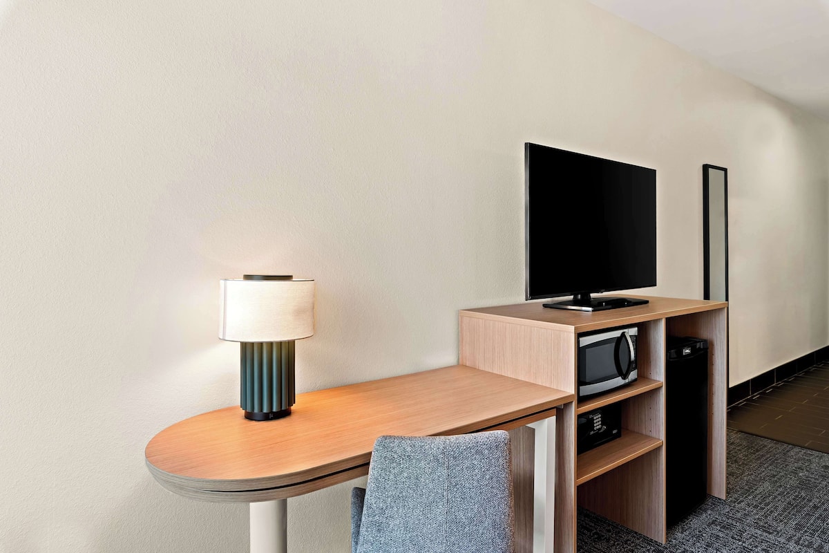 A compact hotel room with a wooden desk, a flat-screen TV, a microwave, and a small shelf.