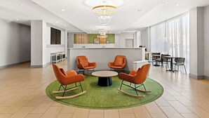 A modern lobby with a reception desk, a circular seating area with orange chairs, a chandelier, and large windows.