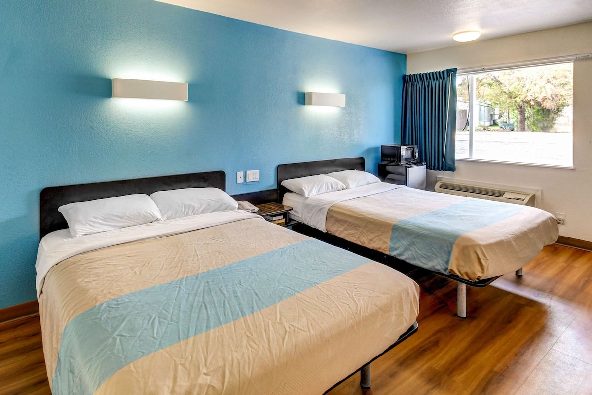 A hotel room with two beds, a window with curtains, a heater, and wall-mounted lights.