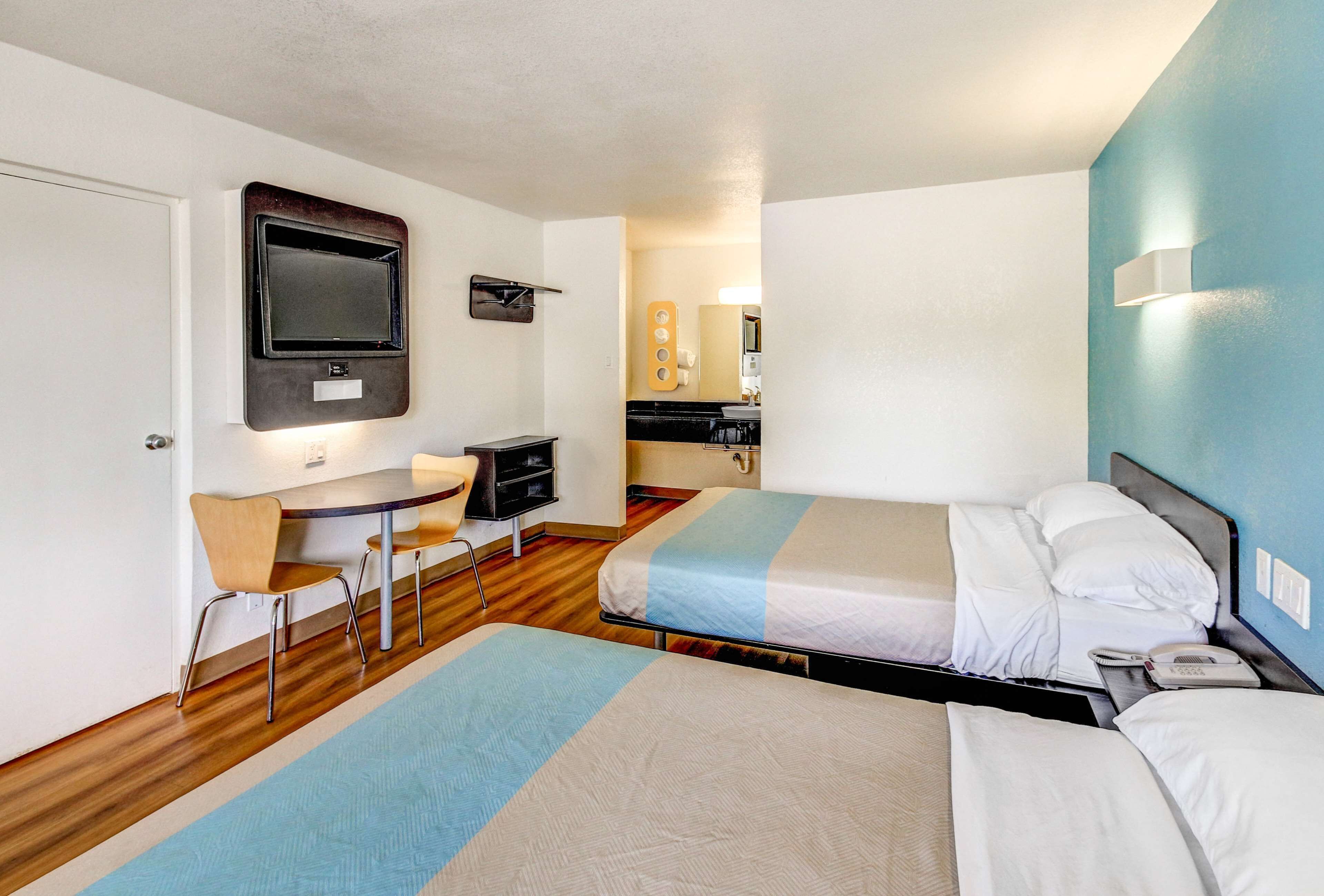 Habitación de hotel con dos camas, un escritorio con silla, un televisor montado en la pared y una pequeña kitchenette.