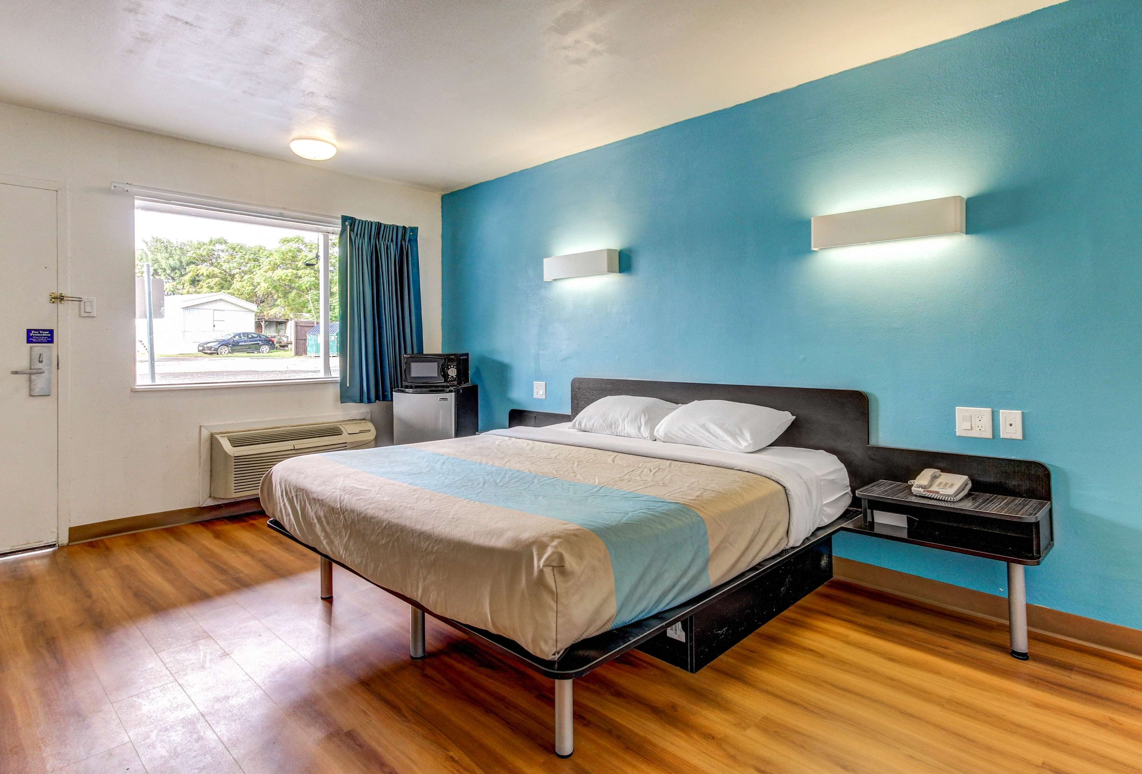 A hotel room with a bed, a bedside table with a phone, a heater, a window with curtains, and a view of the outside.