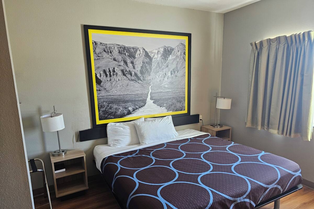 A hotel room with a bed, a nightstand, a lamp, a framed picture of a landscape, and a window with curtains.