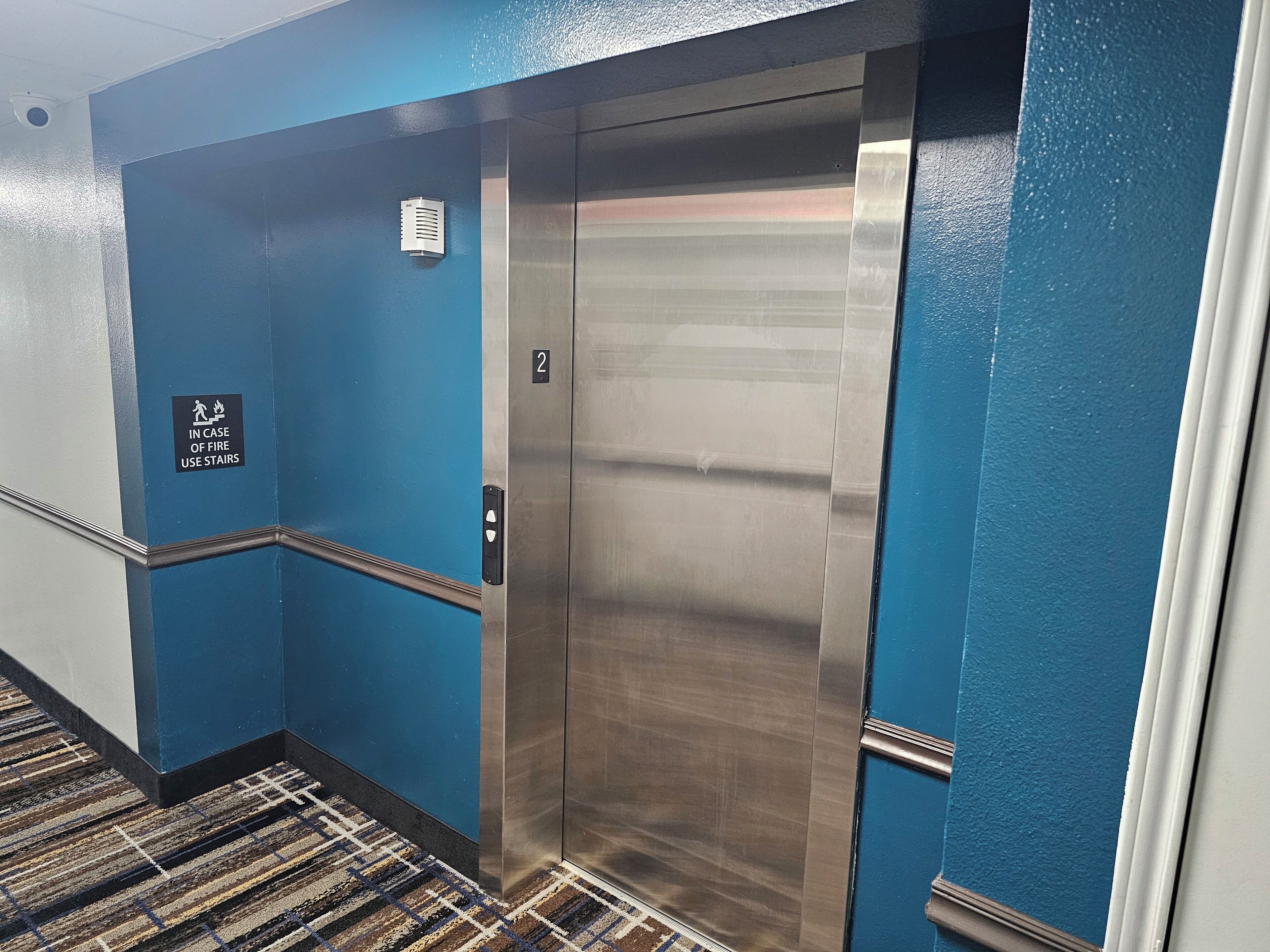 Un ascensor con detalles metálicos, paredes azules y un letrero que indica el uso de las escaleras en caso de incendio.
