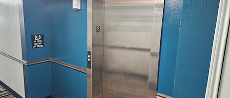 Un ascensor con detalles metálicos, paredes azules y un letrero que indica el uso de las escaleras en caso de incendio.