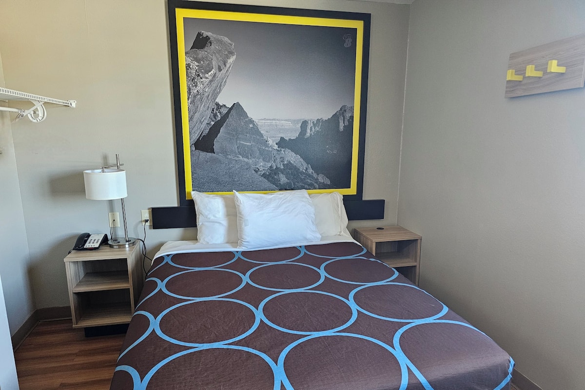 A hotel room with a bed, a nightstand, a lamp, a phone, and a framed picture on the wall.