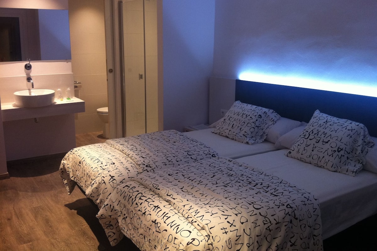 Un dormitorio con una cama, dos almohadas con diseños alfabéticos, una mesita de noche, un espejo y un baño con lavamanos e inodoro.
