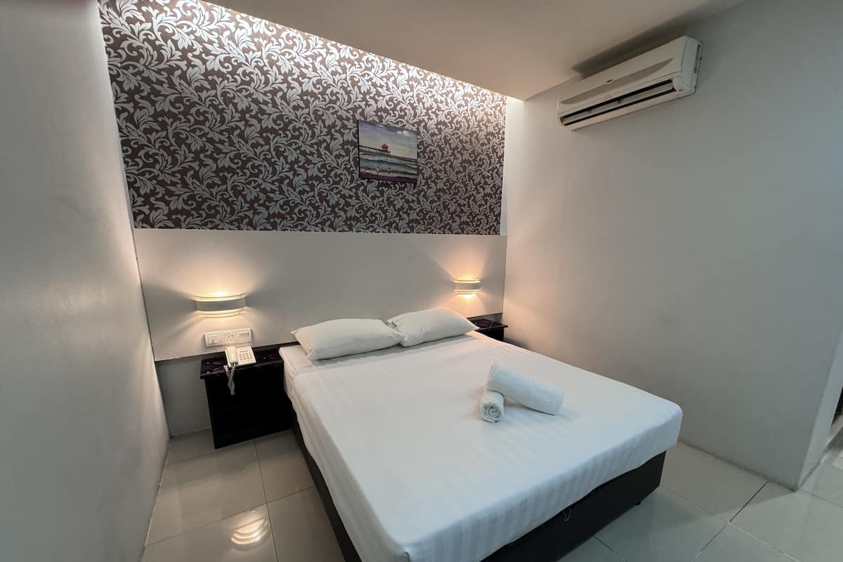 A neatly made bed with a rolled towel, two bedside lamps, a small table with a phone, and a wall-mounted air conditioner.