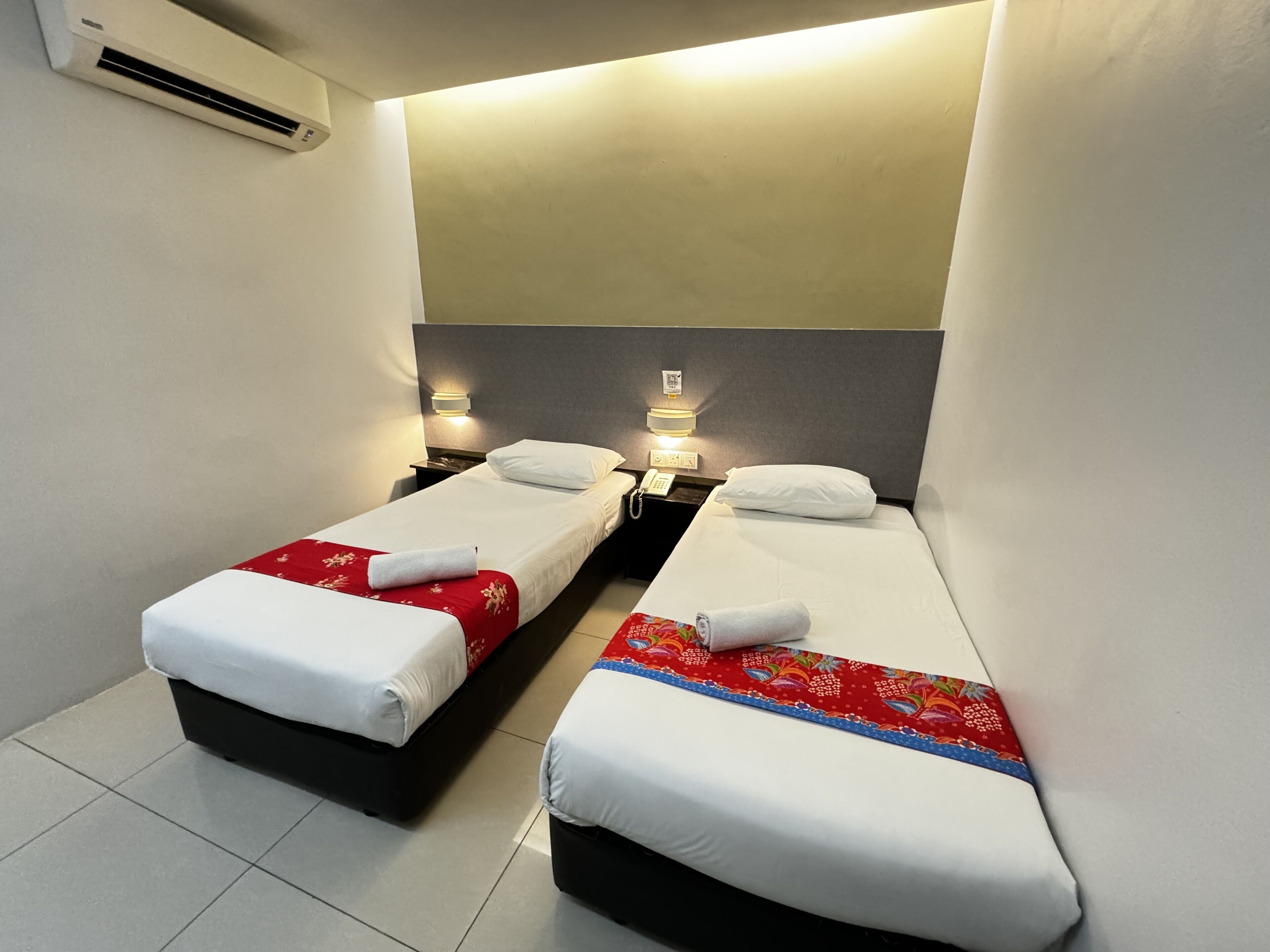 A hotel room with two single beds, each with a red and white bedspread, white pillows, and a rolled towel. There are bedside tables with lamps, a wall-mounted air conditioner, and a ceiling light.