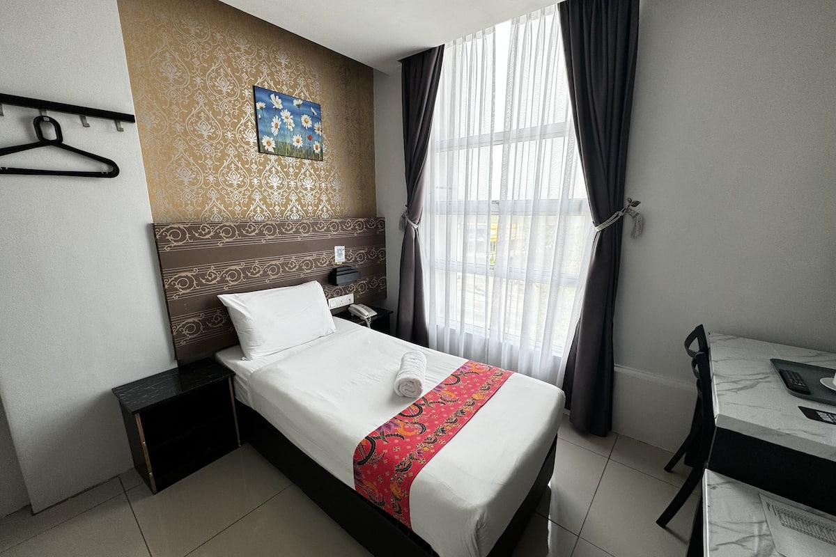 A hotel room with a bed, a desk, a chair, a nightstand, a wall clock, a painting, and a window with curtains.