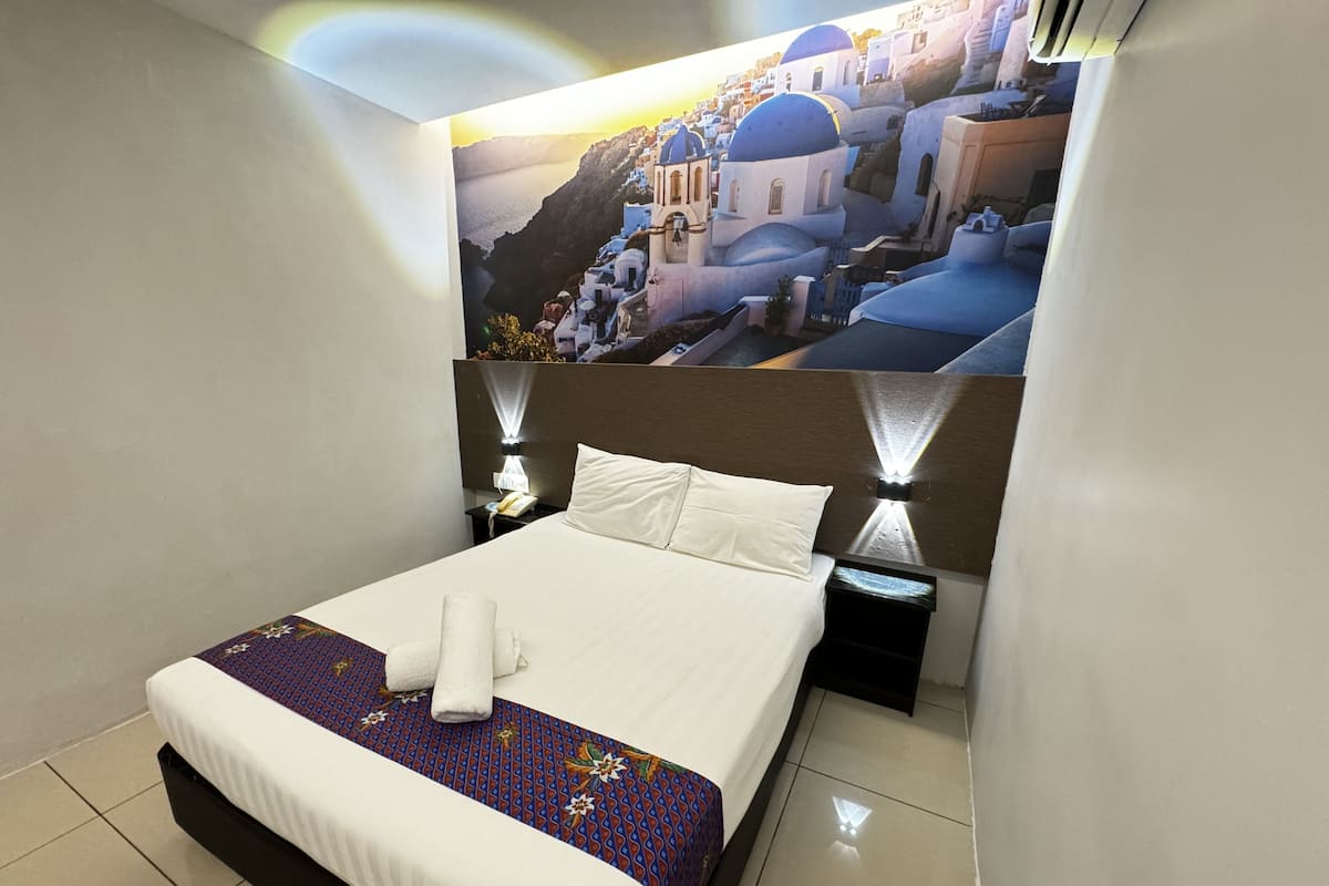 A hotel room with a bed, a nightstand, a wall-mounted lamp, and a poster of a coastal town.
