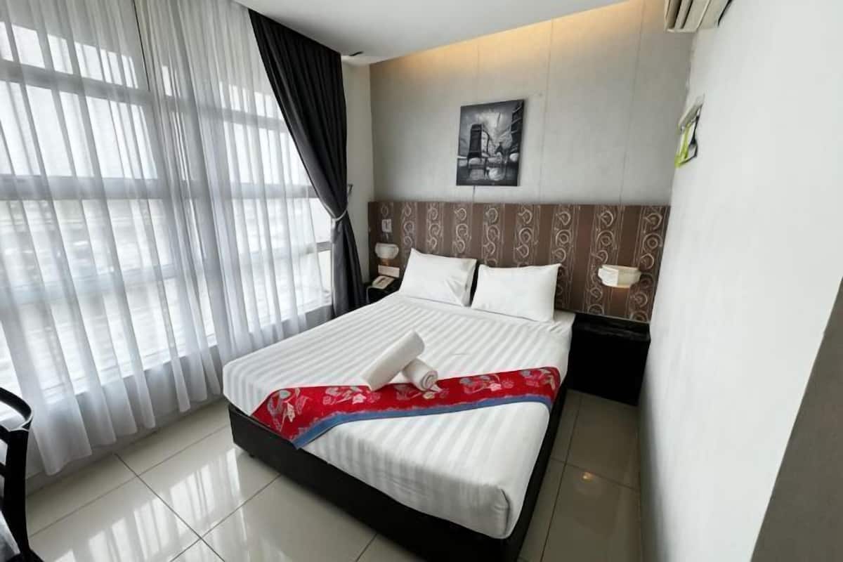 A hotel room with a bed, a nightstand, a wall-mounted lamp, a window with sheer curtains, and a framed picture on the wall.