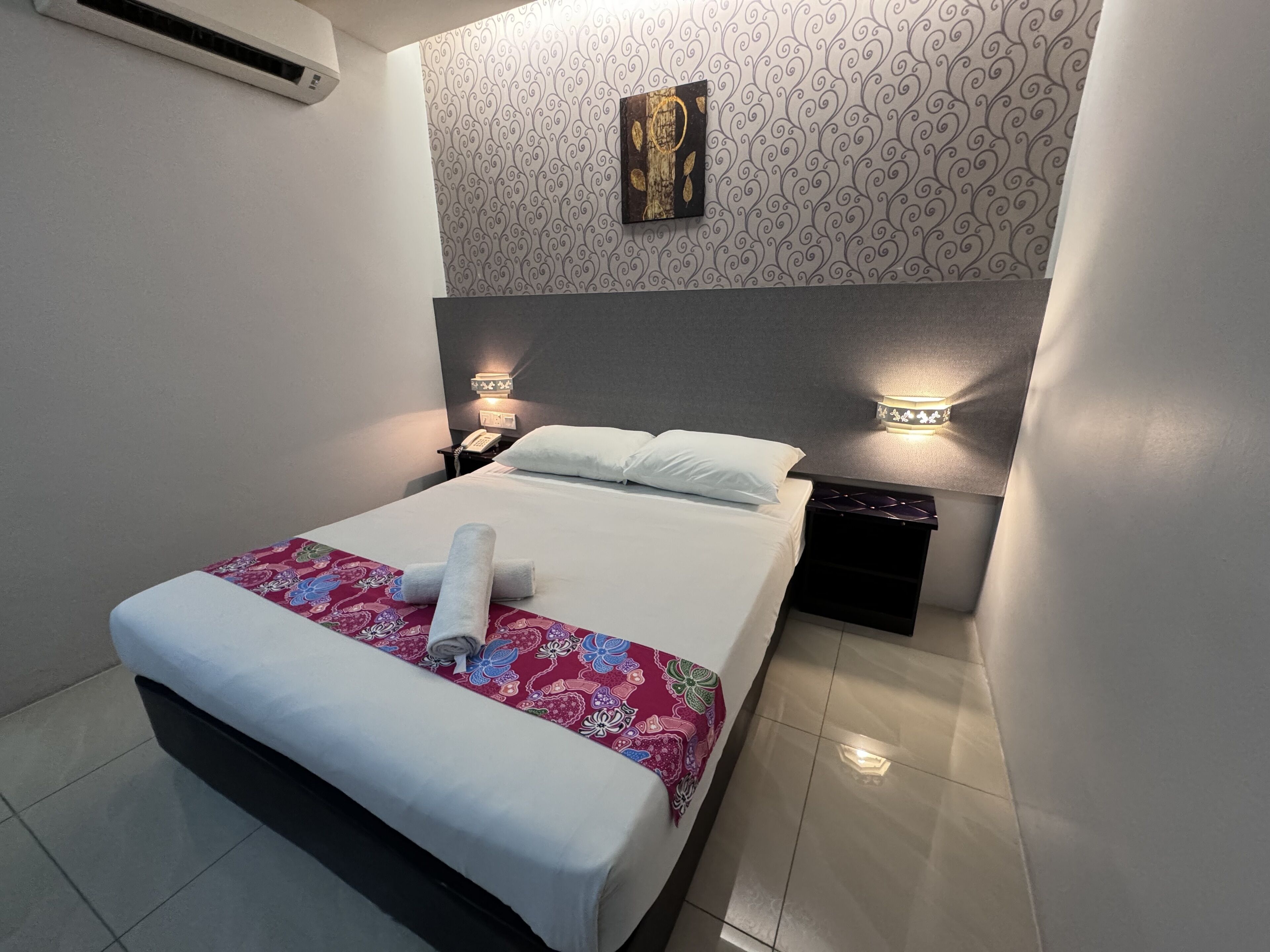 A neatly made bed with a rolled towel, a patterned throw, and a wall-mounted artwork above it.