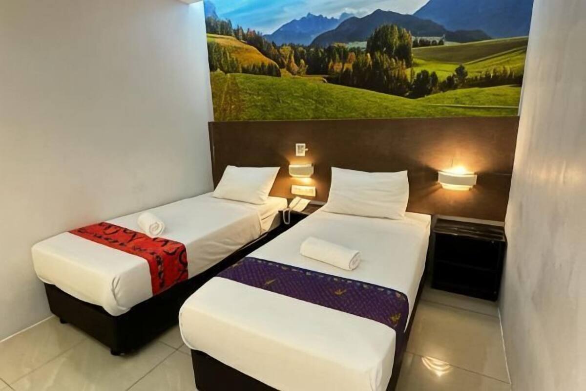 A hotel room with two beds, a nightstand, a wall-mounted lamp, and a scenic landscape mural.