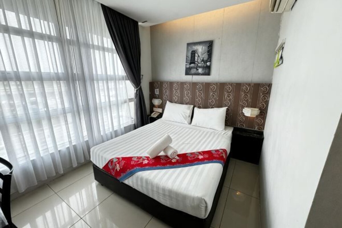 Une chambre d’hôtel avec un lit, une table de chevet, une lampe fixée au mur, une fenêtre avec des rideaux légers et un tableau encadré accroché au mur.