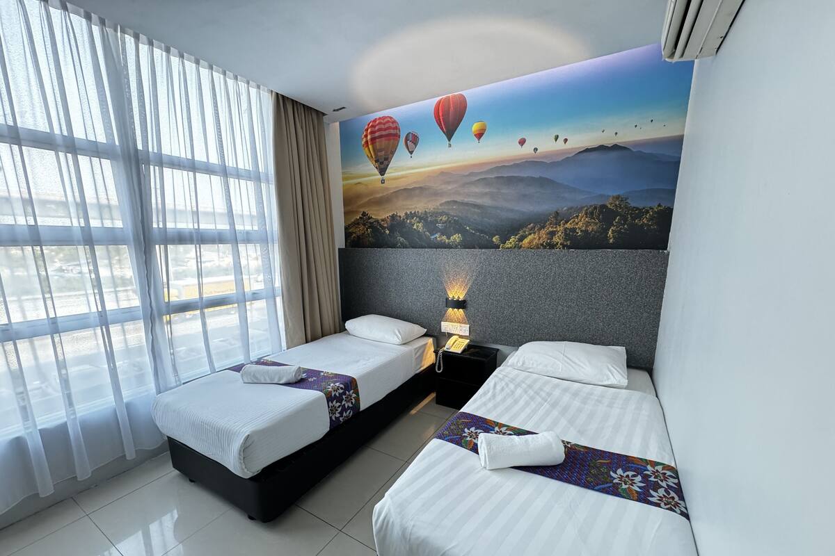 A hotel room with two beds, a large window with curtains, a wall mural of hot air balloons, and a nightstand with a lamp.