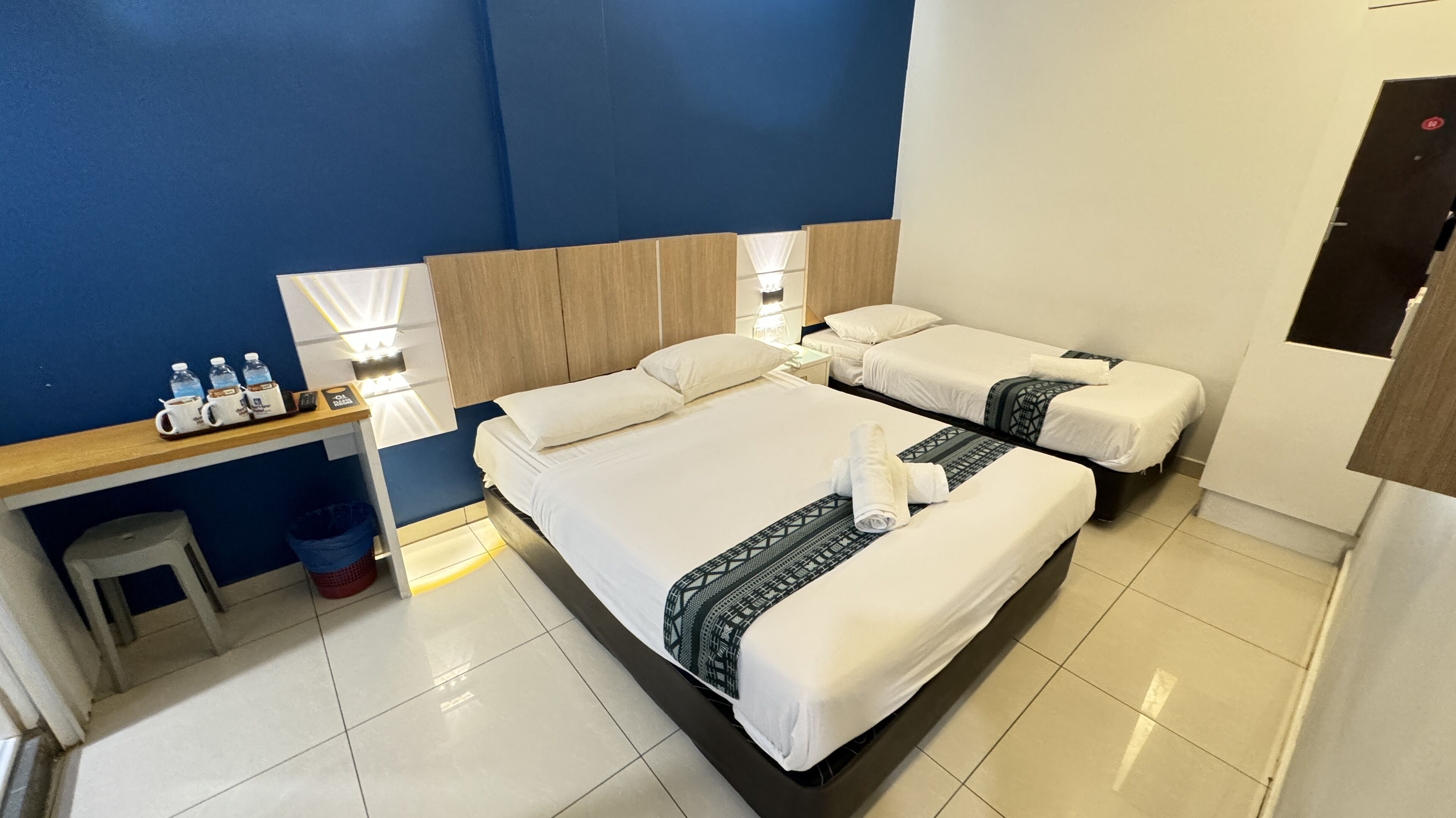 A hotel room with two beds, a desk with water bottles, a trash bin, and a mirror.