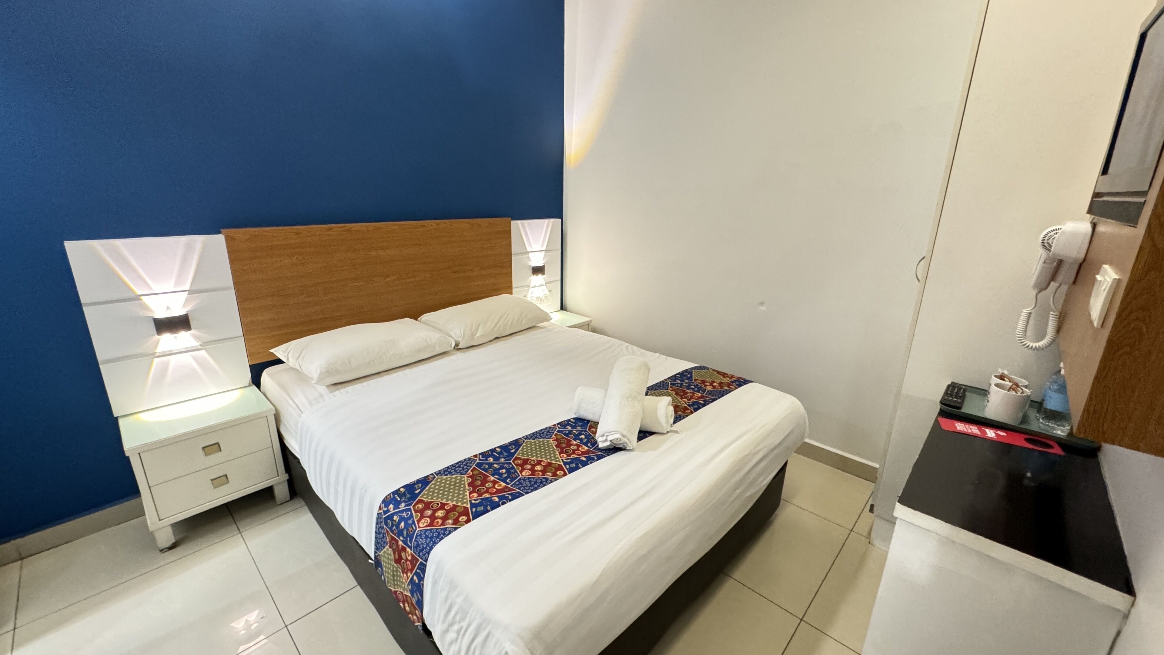 A hotel room with a bed, bedside tables, a nightstand, a desk with a phone, and a mirror.