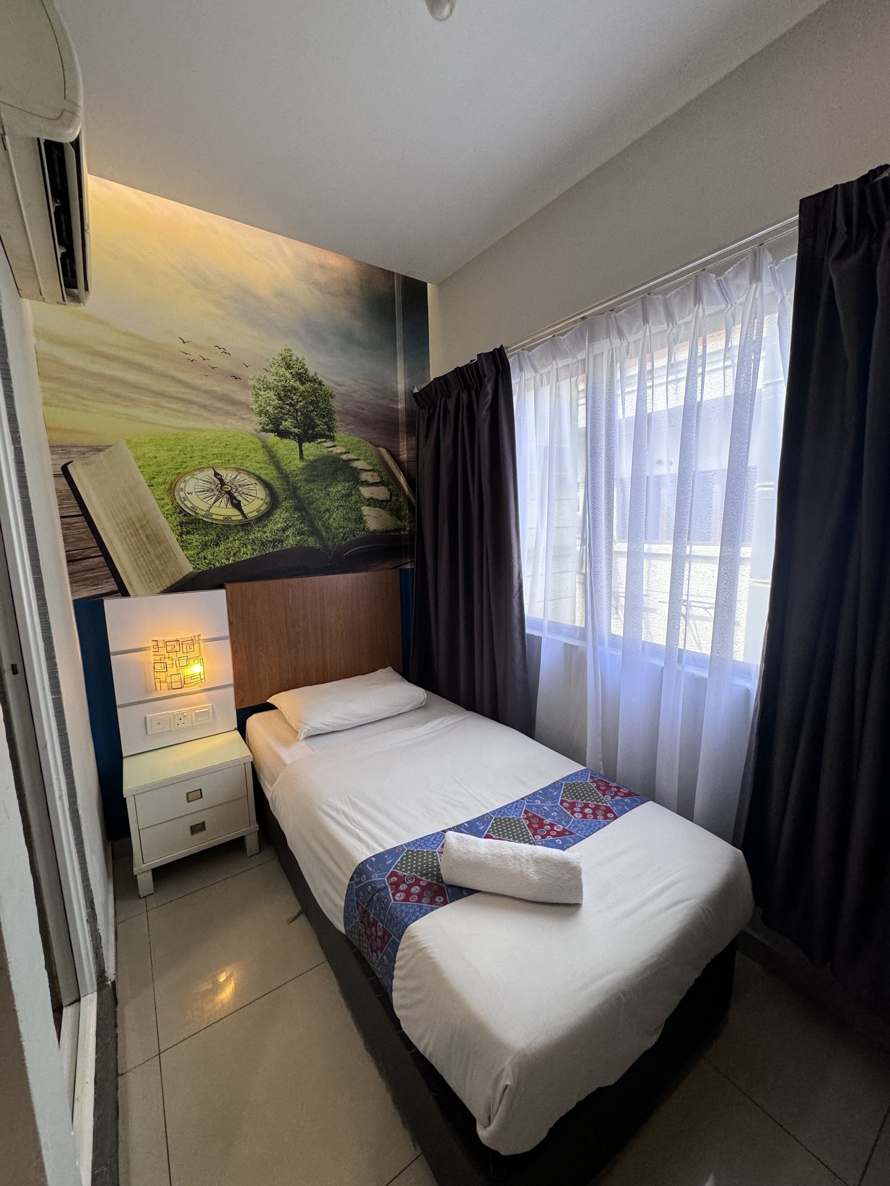 A hotel room with a bed, a nightstand, a wall-mounted lamp, a window with curtains, and a mural on the wall.