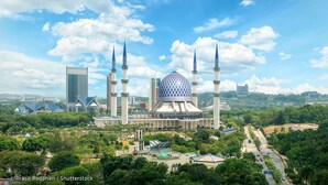 A mosque with a large central dome and four minarets, surrounded by lush greenery and modern buildings.