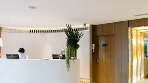 A modern reception desk with a plant, a person seated behind it, and a wooden door to the right. - Citrus Sukhumvit 11 Bangkok by Compass Hospitality (Bangkok)